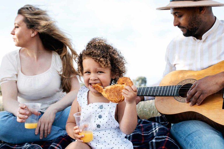 delighted-multiracial-family-enjoying-picnic-together-while-eating-snacks-and-playing-guitar-in-nature-ADSF28352.jpg